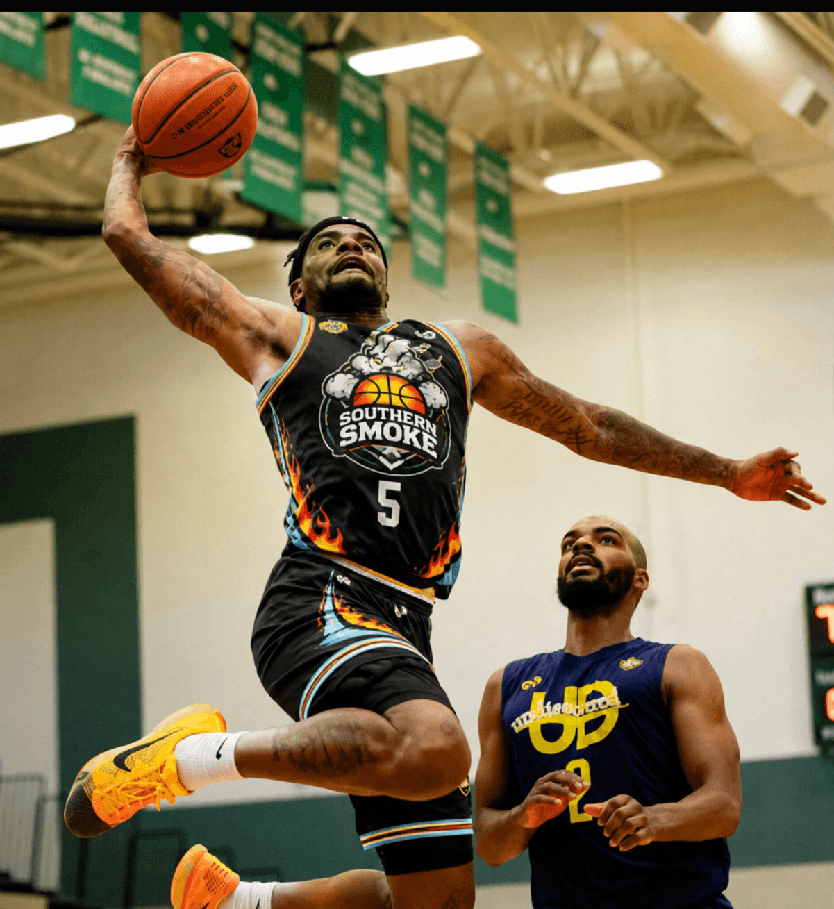 Tattooed basketball player in a black jersey performs a powerful one-handed dunk in a gym.